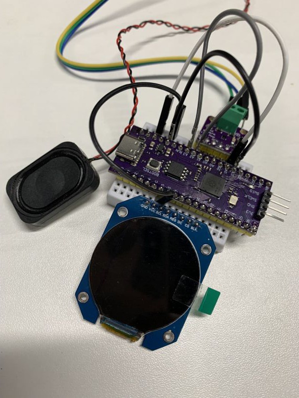 A prototype device built on a small breadboard, with a Raspberry Pi Pico development board, a round display, and an audio amplifier module connected to a small speaker.